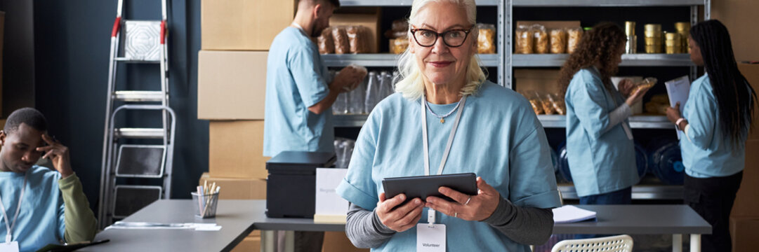 Elderly woman holding tablet and managing inventory at food bank while other volunteers organizing and sorting food items in background. Visible cooperation among volunteers