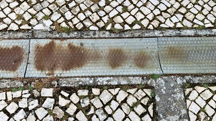 Rust-touched drain amidst cobblestones, reminiscent of forgotten urban labyrinths, evokes Dia de...