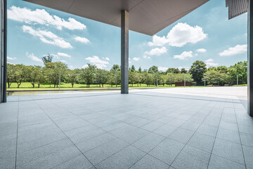 Empty tiled plaza floor providing a clean foreground, with a modern canopy structure above and lush green park trees under blue sky.