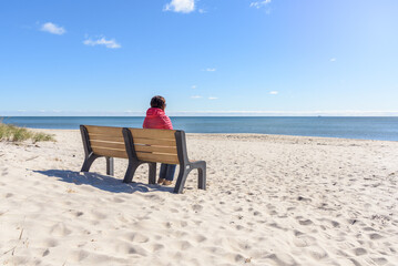 Woman sitting alone on a bench facing a large sandy beach and the ocean on a sunny autumn day
