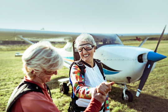 A senior lesbian couple stands on a field, airplane in the backdrop, beaming with pride and exhilaration after skydiving. Their smiles hint at a bucket list dream fulfilled.