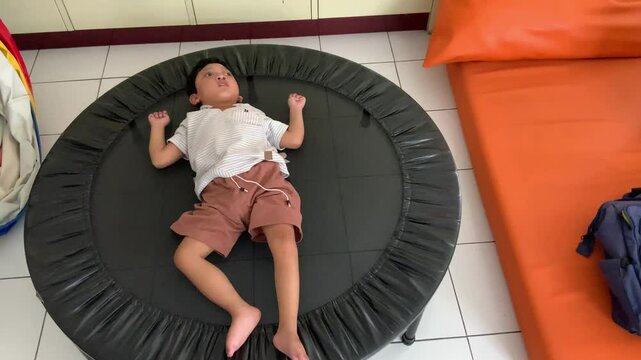 A young boy lies on a black trampoline, his arms and legs spread out, appearing relaxed and peaceful, possibly after jumping