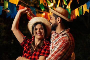 Smiling couple dancing at Brazilian Festa Junina. Both wear traditional plaid shirts and straw hats, with colorful flags and lights glowing in the background