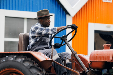 African American young man tractor driver preparing to harvest wheat, hay in rainy weather against background of mill