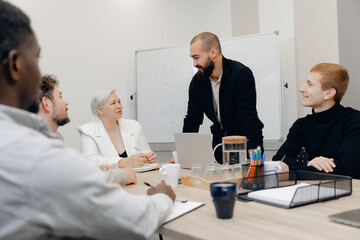 Diverse team in modern office engaged in discussion led by bearded man standing by whiteboard. drinks, stationery, and laptop create professional setting focused on planning