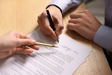 Woman pointing at document and man putting signature at wooden table, closeup