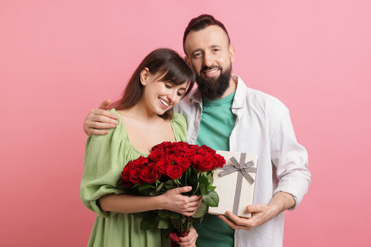 Lovely couple with bouquet of red roses and gift box on pink background