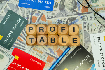 Some hundred dollar bills, some credit cards, a blue pen and a wooden block that says "PROFITABLE". they are all arranged on a work desk. Business and Finance concept