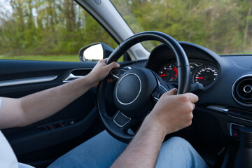 A person is driving a car, gripping the steering wheel with both hands. The dashboard and side...