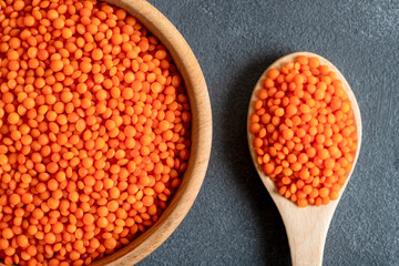 Red lentil grains in a wooden bowl and spoon on a gray background. Top view of dried orange lentils inside the bow
