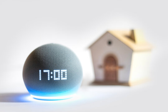 Dispositivo Alexa Echo Dot mostrando la hora con luz LED azul activada y figura de casita desenfocada al fondo, simbolizando un hogar inteligente.