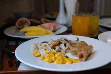 on a white plate lunch dinner corn rice vermicelli with soy meat and two fried spring rolls mango and passion fruit juice as well as fruits for breakfast at the hotel buffet. High quality photo
