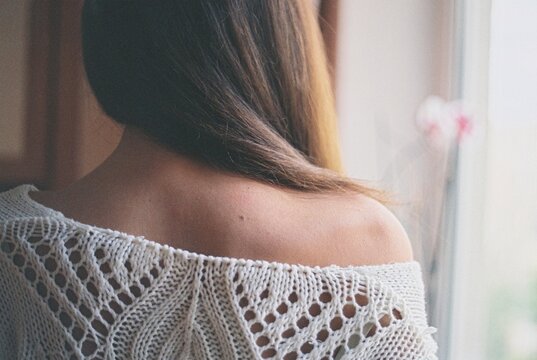 Rear view of a woman with long hair standing by a window wearing on off the shoulder jumper