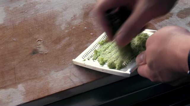 Grating fresh wasabi root on small ceramic grater, hands preparing spicy condiment in kitchen, Japanese cooking ingredient, traditional food preparation, culinary detail