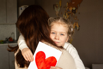 Little daughter with golden hair presents hand-drawn card to her smiling mother. Genuine moment of...