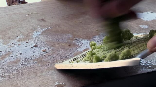 Grating fresh wasabi root on traditional ceramic grater, preparing pungent green paste in kitchen, Japanese cooking, hands working on wooden board, food preparation