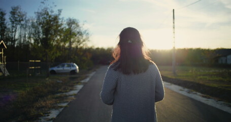 Woman walking down a quiet road at sunset, back turned, peaceful countryside atmosphere, car parked...