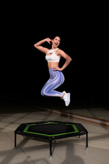 Energetic young fitness girl in blue iridescent leggings and white top jumping and posing on mobile mini trampoline. Jumping workout. High intensity cardio workout, women's health, fun exercise