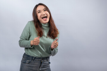 Fototapeta premium Asian woman in green turtleneck smiling cheerfully and giving double thumbs up while winking, against a plain light background