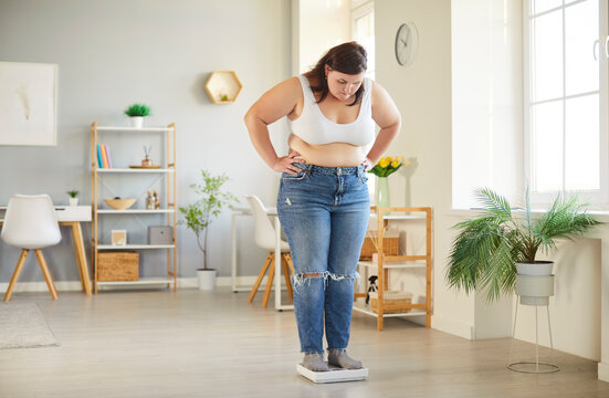Young fat overweight woman in casual clothes looking on the scales at home. Plump plus size girl measuring her weight standing indoors. Obesity, weight control and slimming concept.