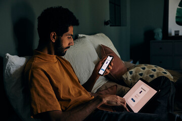 Man typing authentication password on laptop from smart phone while sitting in bedroom at home