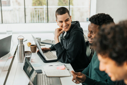 Happy non-binary software engineer sitting at desk while talking with colleague  in tech startup company