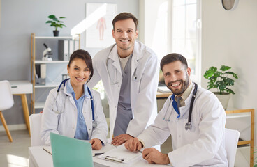 Group of smiling doctors in white coats looking at camera gathered by desk in medical office. Happy professional team of healthcare workers working together in hospital, representing medical field.