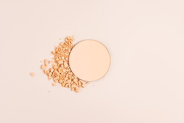 A round block of compact powder in a natural tone and dry broken pieces of dark powder on a beige...
