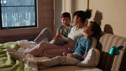 A Black adult woman sitting on a bed, hugging her two school-age sons while talking to them and giving them high fives in a cozy bedroom with a scenic outdoor view.