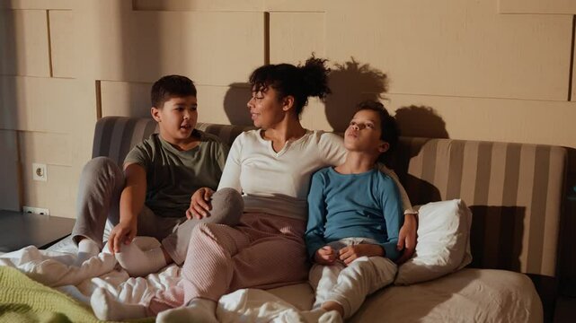 A Latina mother in her 30s with dark hair tied in a ponytail sitting on a bed and talking with two school-aged sons with dark hair dressed in home clothes in a cozy bedroom with beige walls