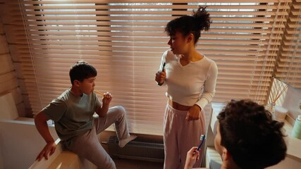 Mother dancing with two sons standing and sitting next to her, they are holding toothbrushes, in the bathroom