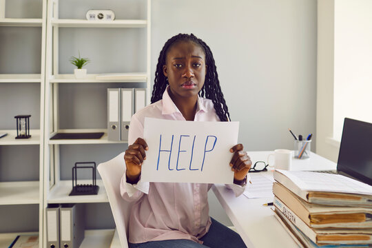 African American woman office worker sits at desk with paperwork and laptop, looks at camera with sad face expression, shows HELP sign, feels stuck at work with unequal pay and no career perspectives