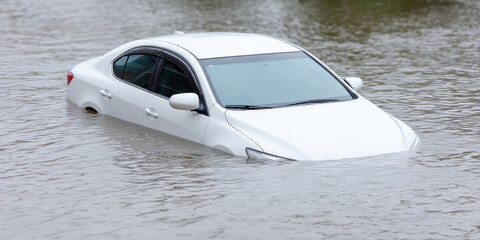 A white car is partially submerged in floodwaters, with water covering more than half of its height. The image concept is natural disaster and flooding