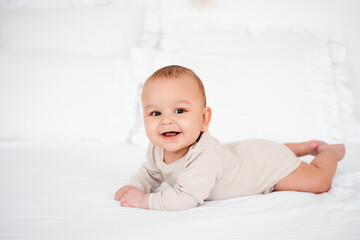a baby, a smiling baby on a white bed on his tummy at home, a healthy, joyful little baby boy in a...