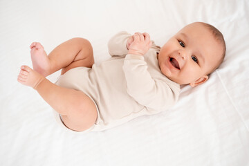 A baby, smiling and joyful baby on a white bed, top view. Healthy, small child boy in beige bodysuit