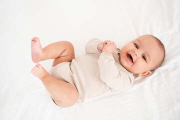 A baby, smiling and joyful baby on a white bed, top view. Healthy, small child boy in beige bodysuit