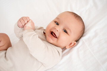 A baby, smiling and joyful baby on a white bed, top view. Healthy, small child boy in beige bodysuit