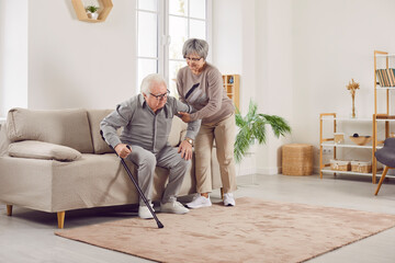 Elderly couple at home, senior wife supports husband to stand with cane for walk from the sofa for rehabilitation after a stroke. Physiotherapy routine, showing care and support in home environment.