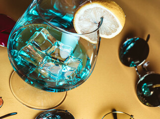 Summer composition with retro-style sunglasses in different colors around a blue cocktail with ice and lemon slices, on a beige background.