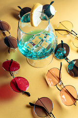 Summer composition with retro-style sunglasses in different colors around a blue cocktail with ice and lemon slices, on a beige background.