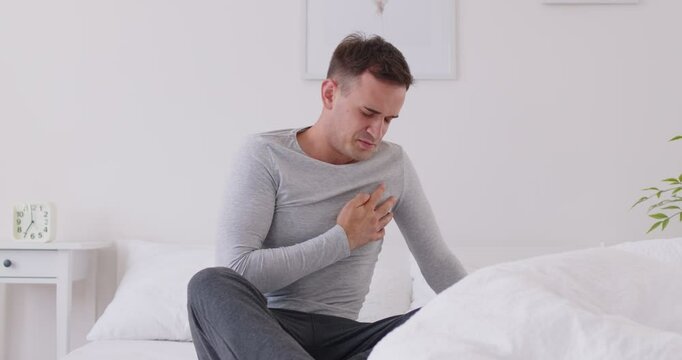 Man having symptoms of a heart attack. Stressed young man in pyjamas sitting on the bed at home in the morning, feeling pain and holding his hand on his chest