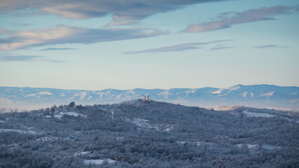 Hilly village covered with frost after cold winter night, there is church on top of hill surrounded with deciduous forest, mountain range in distance