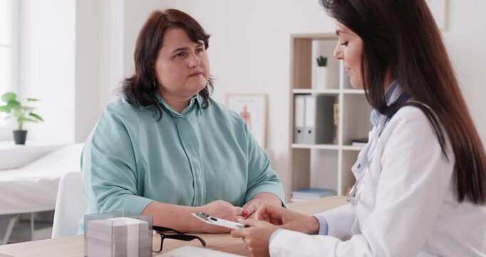 Friendly doctor talks to worried anxious fat chubby ill woman patient with cardiovascular heart disease, gives professional therapy recommendation writes prescription on clipboard, supports, cheers up