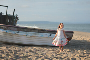 Little beautiful girl in dress stands on the beach near the boat in summer