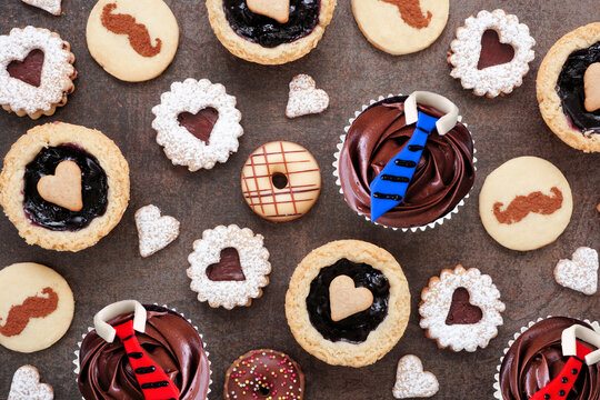 Fathers Day themed baking background with a selection of cookies and sweet treats. Above view over dark stone. Moustache and heart cookies, tarts and shirt and tie cupcakes.