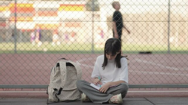 Focused young schoolgirl studies and writes in notebook outside after school near sports ground with students playing.