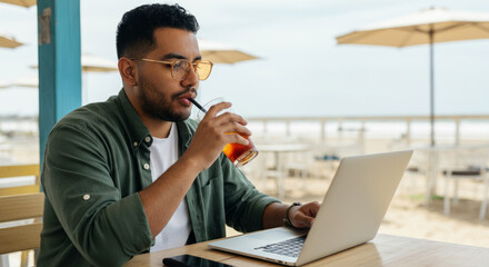 Young hispanic male working on laptop at beachfront cafe