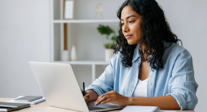 Hispanic young adult female working remotely on laptop in modern home office