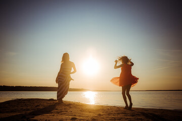 Silhouettes of mother and daughter in dresses jumping and dancing on the beach at sunset. Good...