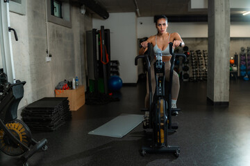 Strong, focused woman powerfully cycles on an air bike during an intense cardio workout,...